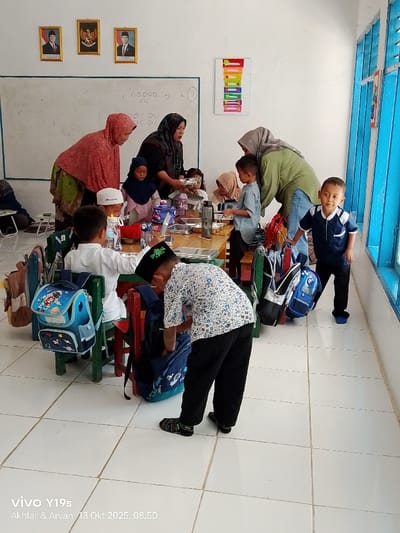 An indoor scene in a classroom or dining area at an educational facility in Jagapura Kulon, Indonesia, depicts approximately three adult women interacting with nine young children during what appears to be a meal or snack time. The room features white walls, a white tiled floor, and bright blue-framed windows on the right side. On the left wall, a whiteboard displays handwritten numbers and symbols, beneath three framed portraits, including the Indonesian Garuda Pancasila emblem. A vertical multi-colored chart is also affixed to the wall.
Three adult women, all wearing hijabs, are present. One woman in a reddish-pink hijab leans over a wooden table, smiling at a child. Another woman in a dark patterned hijab serves food from a large metal tray into individual smaller metal bento-style trays for the children seated at the table. A third woman in a light olive green hijab stands near the blue windows, observing the children.
Several children, primarily boys, are visible. Multiple children are seated at small wooden tables with colorful chairs, some with individual meal trays and water bottles. One boy in a white collared shirt and skullcap sits at a table. Another boy in a patterned white shirt and black peci bends over, actively looking into a blue backpack resting on a red chair. A third boy, wearing a dark blue collared shirt and black pants, stands near the windows on the far right, looking directly at the camera. Various children's backpacks, including one with an astronaut motif, are hanging on the backs of chairs. The overall activity suggests a supervised meal distribution in a structured environment.