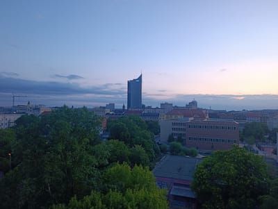 This is a high-angle, wide shot of the Leipzig skyline at dusk. A tall, modern skyscraper with a pointed top, the City-Hochhaus Leipzig, is visible in the center of the frame. The sky is a gradient of blue, transitioning to pink and orange hues near the horizon. Various other buildings, some residential and some commercial, are scattered throughout the city, with some construction cranes visible in the distance. In the foreground, dense green trees obscure parts of the cityscape, suggesting a park or residential area with abundant foliage. The overall atmosphere is calm and serene, characteristic of early evening.