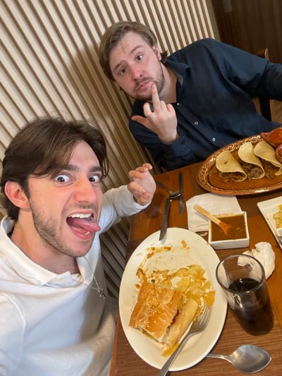 Two men are seated at a table in what appears to be a restaurant. The man in the foreground is looking at the camera with his mouth open and tongue sticking out. He is wearing a white collared shirt and a silver necklace. The man in the background is wearing a dark collared shirt and is giving a middle finger to the camera. In front of them are plates of food, including tacos and a sandwich, as well as glasses of dark liquid and silverware. The table is set on a woven placemat. The background wall is made of vertical wooden slats.