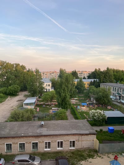 The image presents an elevated view of a residential area in Shumerlya, Russia. In the foreground, a brick building with a flat grey roof is positioned alongside a light grey sedan car. The building has symmetrically arranged windows with dark green frames. Adjacent to the building is a playground with a variety of play structures, surrounded by green grass and trees. In the background, multiple residential buildings of varying heights and colors are visible. A light blue and pink sky with contrails forms the backdrop. The scene appears quiet and devoid of human activity.