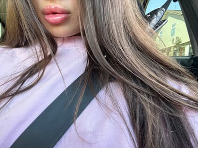 An extreme close-up shot focuses on a person's lips and part of their face, framed by their long, straight, brown hair. The lips are plump and shiny, with a pink, glossy finish. The person is wearing a light pink, high-necked sweater, over which a dark gray seatbelt is diagonally positioned. The background is blurred but suggests an outdoor environment with a building and sky visible through a window, potentially from inside a vehicle. The overall impression is a personal selfie taken in transit. The location is identified as Basarabeasca, Moldova.
