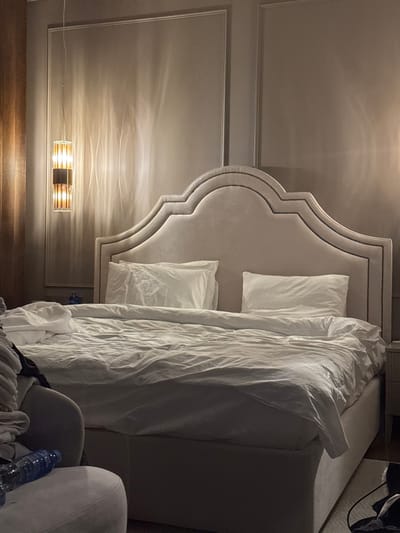 A horizontally oriented shot reveals a mostly made bed with a large, upholstered headboard. Two white pillows rest beneath a rumpled white duvet. A modern sconce mounted on the wall provides illumination. The headboard is lightly colored and stands out against the darker wall. A water bottle and clothing are visible on a chair in the foreground, slightly obscuring the view of the bottom right corner of the bed.
