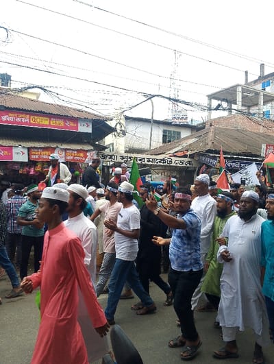 A large group of men, mostly wearing white shirts and pants, walk down a street in Bhola, Bangladesh. The men are all wearing white skullcaps and many are carrying green flags with white text. Some of the men are carrying mobile phones. The scene is set in a densely populated area, with many buildings and electrical wires overhead. The street appears to be narrow and congested. There is a motorcycle parked on the left side of the street. The men are walking in the direction of the camera. The men appear to be participating in a religious procession. The environment is relatively calm and orderly, with no signs of violence or unrest. The location of the image is a street in the city of Bhola, Bangladesh, at coordinates 22.685° N, 90.644° E.