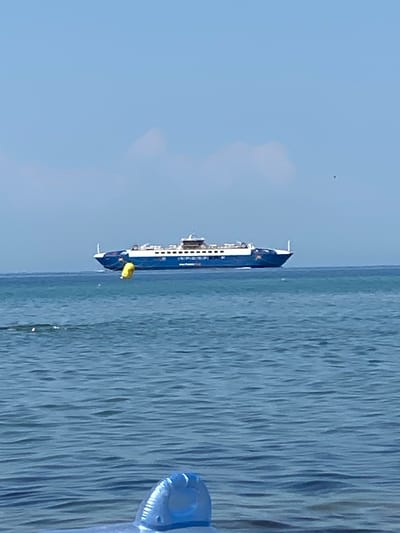 A wide, bright blue ferry with white accents sails across a calm, blue ocean under a clear, pale blue sky. The ferry is positioned in the middle ground of the image, moving from left to right. A bright yellow buoy is visible in the water to the left of the ferry. In the foreground, out of focus, a blue inflatable object is partially submerged in the water, suggesting a beach or recreational area. The location is identified as Keramoti, Greece.