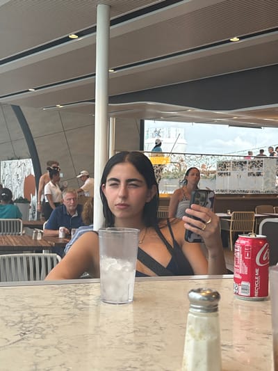 The image depicts a contemporary dining or bar area, located in the city of Falmouth, Jamaica, offering a view of a port. In the immediate foreground, a young adult female with dark hair, wearing a dark-colored top, is seated, looking towards the left with her eyes partially closed. Her left hand holds a smartphone with a camouflage-patterned case; a gold ring is visible on her left ring finger, and a small tattoo is present on her left forearm. On the light-colored, patterned countertop in front of her are a clear plastic cup filled with ice and liquid, a red Coca-Cola can, and a silver-topped shaker.

In the mid-ground, behind the primary subject, another female is seated or standing, looking left. Further back, an older male with gray hair, wearing a blue polo shirt, is seated at a table with a white beverage can. Other individuals, including one possibly identified as staff in a white shirt and another bare-chested male with sunglasses, are observed standing or moving. The area features modern architecture, including a prominent white structural column, light-colored tables and chairs, and a segmented ceiling with integrated lighting. A wall with a black and white mural is visible on the far left.

The background reveals an expansive outdoor view through a large opening. A significant white cruise ship is docked or underway in the distance, with visible figures on its upper deck. The sky is overcast. A decorative metal railing separates the foreground interior from the distant outdoor scene.