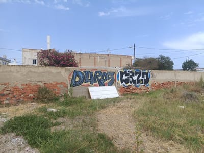 The image depicts a section of a brick and concrete wall, adorned with graffiti, under a clear blue sky with sparse clouds. The wall is located in Almardà, Spain, as indicated by the context. On the wall, two distinct graffiti tags are visible. The one on the left features blue lettering with yellow and black accents, appearing to spell "DUROS" with additional embellishments. To its right, another tag is rendered in black and white, with bubble-style lettering.
In front of the wall, a white, corrugated panel leans against it. The ground in front of the wall is covered with dry grass, weeds, and some patches of green vegetation, suggesting an unkempt or undeveloped area. A few scattered small stones and a larger concrete slab are also present on the ground.
Behind the wall, the upper portions of buildings are visible. One building has a beige facade with visible brickwork and a white chimney or ventilation pipe. A flowering tree with pink blossoms stands prominently in front of this building. Further in the background, more buildings and trees can be seen, along with power lines and streetlights, indicating an urban or semi-urban environment. The overall scene suggests an outdoor, possibly abandoned or industrial area, common in certain parts of Spanish cities. There are no people visible in the image.