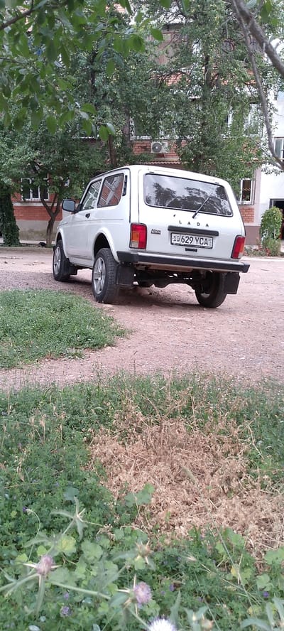 A white Lada Niva is parked on a dirt road in front of a multi-story brick building in the city of Angren, Uzbekistan. The vehicle is facing away from the camera, with its rear bumper and license plate clearly visible. The license plate reads "10 629 YCA". The building appears to be residential, with several windows and an air conditioning unit visible on the exterior. Trees with green foliage surround the building and the parking area. The foreground of the image is dominated by lush green grass and some dry vegetation. The overall scene depicts a typical urban streetscape in Angren.