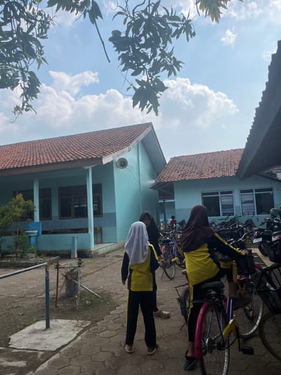 Outside a light-blue, two-story building with a red-tiled roof in Paweden, Indonesia, two individuals stand near bicycles and motorcycles parked on a paved surface. One person wears a brown hijab, a yellow and black uniform, and rides a bicycle. The other person wears a white hijab and a yellow and black uniform. A tree with green leaves is visible in the foreground, and a cloudy sky is overhead.