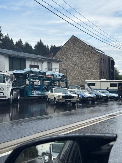 This image captures a street scene in Stavelot, Belgium, during overcast weather, with a wet, reflective paved surface indicating recent precipitation. The foreground features the partial view of a vehicle's black side mirror on the lower right, reflecting a wet road and distant traffic.
In the midground, a collection of classic American performance cars is prominently displayed. On the left, a white transport truck with a blue double-decker car hauler trailer, displaying "COBRA POWER" branding, carries two vintage Ford GT40-style race cars on its upper deck: a dark blue one and a light blue one with a prominent white '6' on its side. At least two smaller blue open-top sports cars, resembling Shelby Cobras, are visible on the lower deck.
Adjacent to the car hauler, a white 1960s Ford Mustang, distinguished by blue racing stripes and a slightly ajar hood, is parked facing the viewer. Behind it, a line of three blue Shelby Cobra-style roadsters, each with white racing stripes, are parked nose-to-tail. Further to the right, a large white recreational vehicle (RV) or bus is parked, with a vintage pickup truck partially visible beside it. Two individuals stand near the rear of the RV. Small white display stands are positioned near the Mustang and the car hauler.
The background features two buildings. To the left stands a white, two-story house with a dark roof and a balcony. To its right, a larger multi-story building displays a prominent stone facade on its left side and dark wooden cladding on its right, with a sloped roof fitted with what appear to be solar panels. Dark conifer trees line a hillside behind the structures. Overhead, multiple black power lines stretch horizontally across the frame against an overcast sky with patches of lighter clouds.