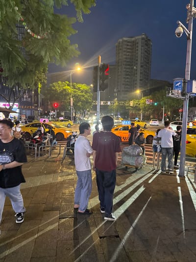 The image depicts a street scene at dusk or night in the city of Longta, China. The area appears to be a busy urban intersection with several yellow taxis visible, indicating a public transportation hub. A tall, multi-story building dominates the background, suggesting a developed urban environment.
In the foreground, several individuals are gathered on a wet sidewalk. The ground is paved with rectangular tiles, and reflections of lights suggest recent rain. The people are dressed in casual attire. Two young men are standing in the center, looking towards the right. To their left, a woman is pushing a cart laden with large, wrapped packages. Other individuals are scattered around the scene, some appearing to be waiting for transportation or interacting with each other.
Streetlights and traffic lights illuminate the scene, casting long shadows across the ground. The sky is dark, with the twilight ambiance suggesting the transition from day to night. Decorations, possibly for a festival or holiday, are visible hanging from a tree in the upper left portion of the image. A surveillance camera is mounted on a pole on the right side of the frame, typical of urban security infrastructure. The overall impression is one of a lively, active urban area during the evening hours.