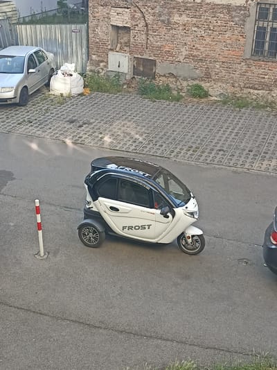 The image depicts a white three-wheeled electric vehicle, branded "FROST" and "ELECTRORIDE", parked on an asphalt road in an urban environment. The vehicle has a black roof and a transparent windshield. To the left of the vehicle, a red and white striped pole is partially visible. In the background, a silver car is parked next to a large white bag filled with debris. Behind the car and the bag, a metal fence and a brick building with a barred window are visible. The ground in front of the fence and building is covered with decorative concrete pavers. The overall scene appears to be a side street or parking area, possibly in Sosnowiec, Poland, given the context provided. There are no people visible in the image.