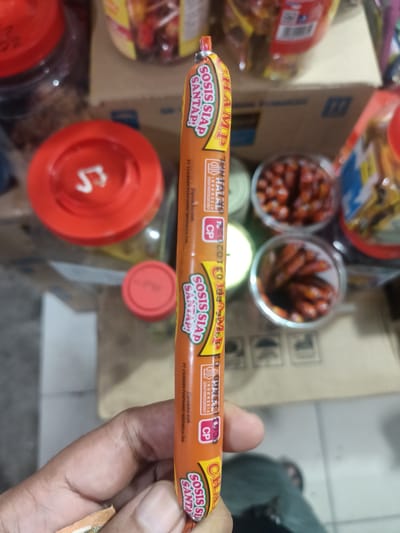 A hand holds a Champ Sosis Siap Santap sausage in its orange packaging. The packaging includes nutritional information, halal certification, and a logo. The background includes food containers, a tiled floor, and a glimpse of a shoe. The image was likely taken in Batam, Indonesia, as indicated on the packaging.