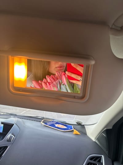 The image depicts the interior of a vehicle, specifically from the passenger's perspective looking towards the driver's side. The overhead sun visor is lowered, revealing an illuminated vanity mirror. Reflected in this mirror is the lower portion of a female's face, primarily her lips and chin, partially obscured by a bouquet of pink tulips wrapped in clear plastic. Her right hand, adorned with a silver ring on the ring finger, is visible holding a red smartphone, suggesting she is taking a selfie using the mirror. An amber-colored light fixture, part of the vanity mirror assembly, is illuminated on the left side of the mirror.
Below the sun visor, the dark gray dashboard of the car is visible, featuring air vents and what appears to be a digital display screen on the left. On the dashboard, a blue fabric face mask with a white design (possibly a sun or eye symbol) and white trim is laid flat. To its right, a small yellow or gold decorative tassel is present. The specific location within the city of Slavyansk-na-Kubani cannot be determined from the provided image beyond the internal setting of a car.