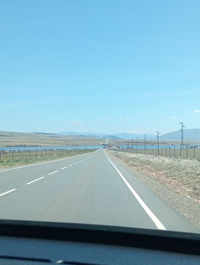The image displays a rural highway scene in Urraca Miguel, Spain, under a clear, light blue sky. The perspective is from a vehicle traveling forward on a two-lane asphalt road, evidenced by the dark dashboard visible at the bottom of the frame. The road features a dashed white center line and a solid white line marking the right shoulder. To the left of the road, a wooden post fence parallels the asphalt, separating it from a calm body of water, likely a reservoir or lake, which extends into the distant landscape. Beyond the water, rolling hills and mountains, some with snow-capped peaks, define the horizon. To the right of the road, a metal fence borders an area of dry, light brown and green vegetation. A series of dark-colored utility poles supporting power lines runs parallel to the road on the right side, receding into the distance. No people or other vehicles are visible.