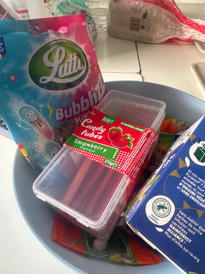 The image displays a collection of candies and other items within a light blue bowl. In the foreground, a clear plastic container holds numerous red, tube-shaped candies, labeled "Candy tubes" with a strawberry flavor. A large, pink and blue bag with the "Lutti" logo and cartoon characters is partially visible to the left. To the right, a blue box with text in French and a Rainforest Alliance certification is present. Various other food items and packaging are scattered around the bowl. The scene appears to be an indoor setting, possibly a kitchen or dining area. There is no indication of the city of Tourcoing, France, or any specific location.