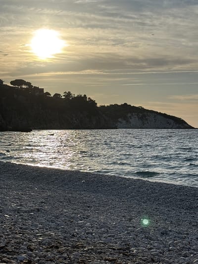 The image displays a serene coastal landscape captured during either sunrise or sunset, set in Portoferraio, Italy. The bright, low-lying sun dominates the left side of the sky, its intense light diffused by soft, wispy clouds. Faint contrails are visible against the pale, warm-toned sky.

To the left, a dark silhouette of a rugged, tree-covered promontory or cliff extends into the frame, its features obscured by the backlighting from the sun. The sea occupies the middle ground, showcasing a dazzling display of sunlight reflecting off its rippled surface, creating shimmering vertical streaks of light.

On the right, a dark pebble or shingle beach stretches into the foreground, with individual stones and shells clearly discernible, indicating a textured surface. A notable detail on the beach is a small, bright green, circular reflection or object visible in the mid-right. No people or visible text are present. The scene evokes a tranquil and atmospheric mood, characteristic of a Mediterranean coastal evening or morning.