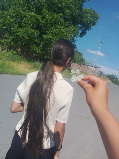 A person is standing on an asphalt road, facing away from the camera. They have very long, dark brown hair tied into a ponytail and are wearing a light-colored, short-sleeved, ribbed shirt and black pants. A hand is visible in the foreground, holding a small cluster of white flowers. In the background, there is a lush green tree, a wire fence, a brick wall, and some buildings. A lamppost and power lines are visible against a bright blue sky with scattered clouds. The scene appears to be in a rural or suburban area, possibly in Nor Geghi, Armenia, given the typical landscape.