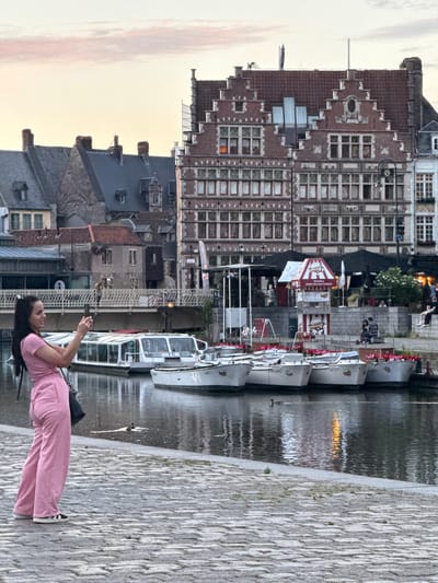 In Ghent, Belgium, a woman in a pink jumpsuit stands on a cobblestone path next to a canal, holding a smartphone to take a picture. She is facing a row of white boats docked on the water. Behind the boats, historic buildings line the canal, with a bridge in the background. The sky is overcast with a hint of sunset. The scene appears to be a tourist area, with several people visible in the distance near outdoor seating areas.