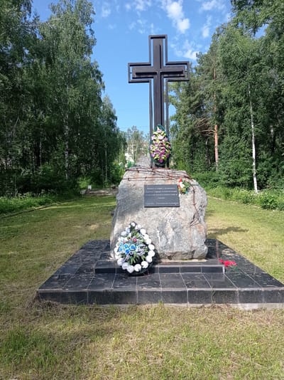 The image shows a memorial with a large black cross atop a boulder. The boulder is surrounded by a black tiled base. Wreaths are placed on the boulder and the base. Text is inscribed on a plaque on the boulder. The memorial is situated in a grassy area with trees in the background. The sky is blue with some clouds. This memorial is located in the city of Degtyarsk, Russia, and is dedicated to the victims of terror and political repression.