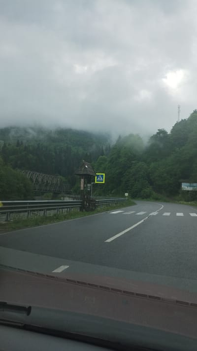 The image depicts a view from inside a vehicle driving on a road in a mountainous, forested area. The scene includes fog partially obscuring the view of the trees and mountains. A pedestrian crossing sign is visible next to a wooden structure resembling a small covered bridge or pavilion. The road is asphalt with white lane markings. Based on the location information provided, the scene is likely in or near the town of Yasinya, Ukraine.