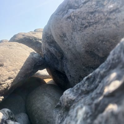 The image shows a close-up view of large, weathered rocks under a clear sky, possibly on a beach or coastline. The rocks vary in color from light brown to dark gray and exhibit textured surfaces. Some rocks are smooth and rounded, while others are more angular. The lighting suggests it is daytime, with sunlight casting shadows across the stones. The scene is devoid of any human presence or activity. The location is identified as Fanore, Ireland, suggesting this is a natural outdoor landscape.