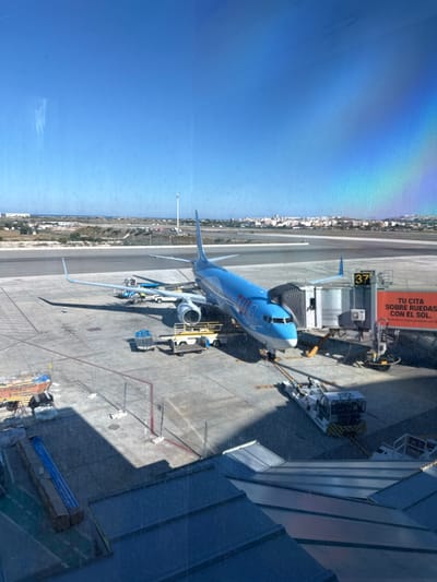 The image depicts an aerial view of an airport scene in Elx / Elche, Spain. A blue and white passenger airplane is docked at gate 37. Support equipment, including a boarding bridge and various service vehicles, surrounds the aircraft. Buildings and structures of the airport terminal and ground facilities are visible in the foreground. The urban landscape of Elx / Elche is seen in the distance, along the coastline. The sky is clear and blue with a rainbow effect in the corner of the photo, possible a reflection in the window.