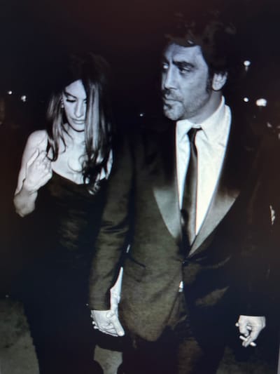 In a dimly lit, outdoor setting, a man and a woman are captured from the waist up, walking hand-in-hand. The man, on the right, is wearing a dark suit with a white shirt and a dark tie. He has short, dark hair and appears to be looking off to the side. He is holding a cigarette in his right hand. The woman, on the left, has long, dark, wavy hair that partially obscures her face. She is wearing a dark, strapless dress and her left hand is resting on her chest, fingers splayed. The background is dark with blurred lights, suggesting an evening or nighttime scene. The location is identified as the city of Protasovo, Russia.