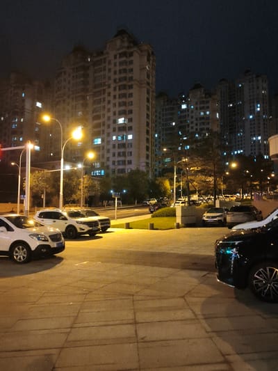 A nighttime street scene in Jixi, China, shows several parked cars on a paved area illuminated by streetlights. In the foreground, a black car is partially visible on the right. To its left, a white SUV is parked, followed by another white SUV and a white sedan. The background features tall apartment buildings with numerous illuminated windows, indicating the presence of residents. Trees are scattered throughout the scene, and streetlights cast a warm glow on the pavement. The sky is dark, characteristic of nighttime.