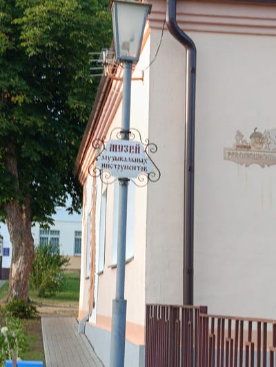 An outdoor medium shot captures a building facade on a sunny day. The building is painted a light cream color with brown trim along the roofline and a dark brown gutter. Attached to a lamppost in front of the building is a decorative sign with Cyrillic text that reads "МУЗЕЙ МУЗЫКАЛЬНЫХ ИНСТРУМЕНТОВ" (Museum of Musical Instruments). To the right of this sign, a smaller decorative plaque with acorn motifs and the year "2023" is visible on the building's wall. A wooden railing with vertical slats runs along the base of the building. To the left, a large tree with green foliage is prominent, and a paved walkway leads towards the building. In the background, another building with white walls and barred windows can be seen. The overall scene suggests a quiet, possibly historic, location in Smilovichi village council, Belarus.