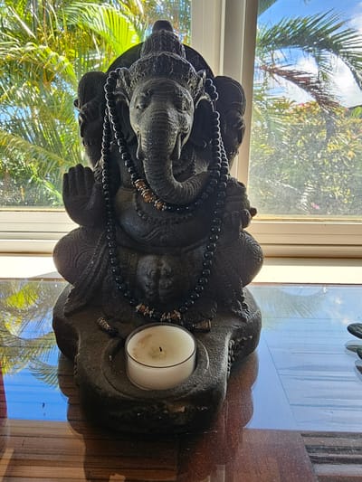An indoor close-up shot depicts a dark stone statue of the Hindu deity Ganesha seated with his hands in a mudra, adorned with black beaded necklaces. The statue is placed on a wooden surface in front of a window with sheer white blinds, through which palm trees and a bright blue sky are visible. A lit white tea candle sits in a stone holder at the base of the statue. The setting is likely Napili, United States of America, given the tropical foliage outside the window.