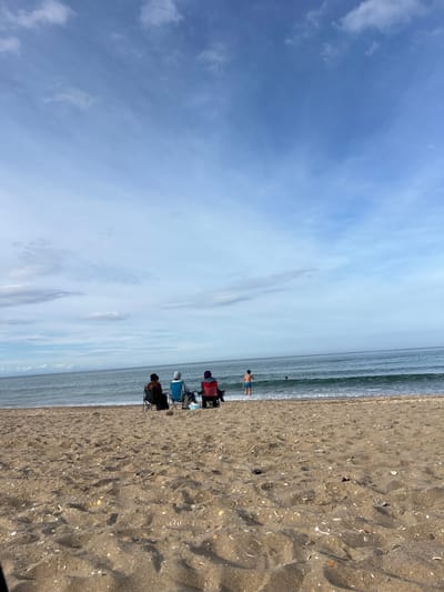 The image depicts a beach in M'Diq, Morocco, on a clear day with a blue sky and scattered clouds. The beach extends along the right side of the frame, covered in sand and seashells. Three beach chairs are positioned near the water's edge, occupied by people. A man is standing in the water, further down the shoreline. The ocean stretches out towards the horizon.