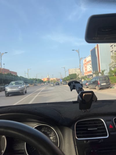 The image shows a view from inside a car driving on a road in Beni Mellal, Morocco. Several cars are on the road, and buildings are visible on either side. Streetlights line the road. A phone mount is attached to the car's dashboard. A rearview mirror and part of the steering wheel are also visible inside the car.