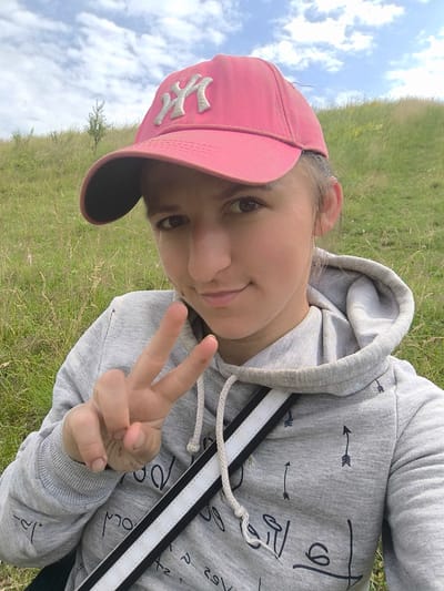 A young woman wearing a pink baseball cap and a grey hoodie is taking a selfie outdoors. She is in a grassy, hilly area, possibly on the outskirts of Anastasivka, Ukraine, given the rural setting and cloudy sky. She is holding up two fingers in a peace sign. The hoodie has a white and black strap across it and a pattern of small black arrows and abstract designs. The background is a blurred hillside covered in dry grass and some greenery, with a few scattered trees and clouds in the sky.