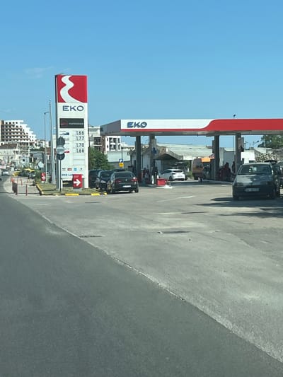 This media file captures a sunny daytime scene at an EKO gas station in Ulcinj - Ulqin, Montenegro. The perspective is from a vehicle, looking towards the gas station and the road.
The primary subjects are the gas station infrastructure, including the prominent red and white EKO branding on the canopy and a tall sign displaying fuel prices. Several cars are visible, some are at the pumps, suggesting the activity of refueling. One dark-colored SUV is parked prominently near the pumps. People are visible under the gas station canopy, appearing to be interacting or waiting.
In the background, there are buildings, suggesting an urban or semi-urban environment. Trees are also present, adding to the outdoor setting. The sky is a clear, bright blue, indicating fair weather. The road surface is visible in the foreground, appearing somewhat worn and textured.
Notable details include the fuel prices displayed on the EKO sign, with numbers such as "166," "177," and "170" visible. The EKO logo, a stylized red "W," is clearly identifiable. The overall impression is of a functional, active gas station on a pleasant day.