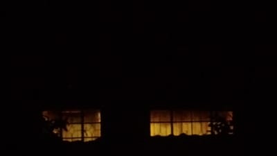 This image captures an exterior, nighttime view of two illuminated windows on a building facade in Ndeiya, Kenya. Both rectangular windows exhibit an internal, diffused orange-yellow light, with visible grilles or bars across their panes. Indistinct dark foliage is present at the immediate base and sides of the windows. The surrounding environment is completely dark, with no other objects, individuals, or actions discernible within the frame.