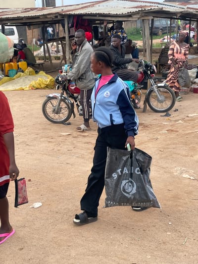 In Wang, Nigeria, a market scene unfolds with several individuals and objects. A young woman in the foreground, wearing a blue and white jacket and black pants, carries a large black shopping bag. She walks from left to right across a dusty, unpaved ground. Behind her, a motorcycle is parked. In the background, a covered market stall is visible, with various goods and people interacting. One man, on his phone, stands near a motorcycle, while another person sits on a bench. The market appears to be a bustling open-air environment with temporary structures.