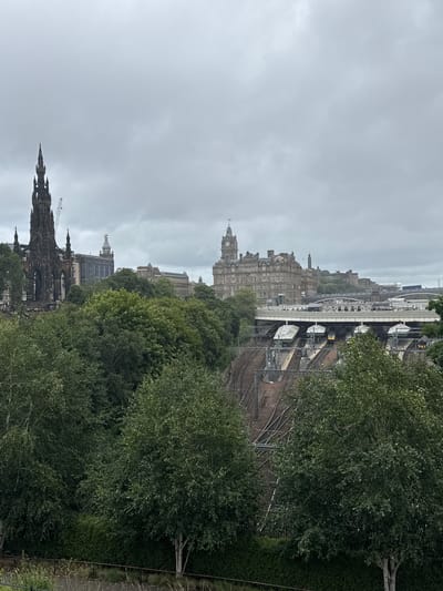 The image depicts an overcast view of a cityscape in Edinburgh, United Kingdom. In the foreground, lush green trees and bushes are visible, with a metal fence and dry grass in the immediate bottom. Behind the foliage, a complex network of railway tracks and platforms is situated, with several trains at the station. Further in the background, several notable buildings and structures are present. To the left, a tall, dark, Gothic-style monument with intricate detailing stands prominently, with a construction crane visible behind it. Adjacent to this monument are other historical buildings. In the center and to the right, a large, multi-story stone building with a clock tower dominates the skyline, appearing to be a significant landmark. Other buildings and bridges are scattered across the horizon, suggesting a dense urban environment. The sky is uniformly grey and cloudy, contributing to a muted color palette of the scene. No people are discernible in the image.
