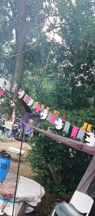 The image depicts an outdoor scene, likely a park or recreational area, with lush green trees forming a canopy. A string of colorful bunting, featuring flags in pink, grey, yellow, and white cutouts of birds or doves, is strung between trees. In the foreground, a large purple flower decoration is prominently displayed, attached to a pole. To the left, a blue plastic bin sits on a table covered with a white and patterned cloth. Several people are visible in the background, engaged in various activities, with some appearing to be walking or standing around. The overall atmosphere suggests a casual outdoor gathering or event. The location is identified as Старые Быги, Russia.