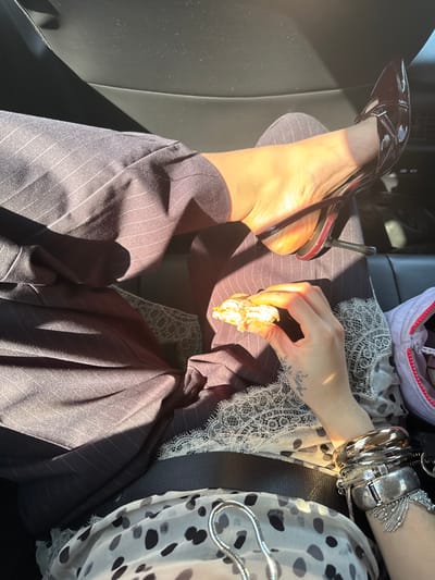 A person in pinstripe trousers and a black patent leather stiletto with red accents is sitting in a car. Their right leg is stretched out, showcasing the shoe. They are holding a small, tan-colored object in their hand. Silver bracelets adorn their wrist. A white lace garment and a polka-dotted top are visible near the waist. The lighting is bright and sunny, casting stark shadows on the leg and trousers.