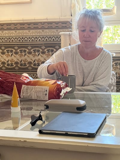 A middle-aged woman with short, light blonde hair is inspecting a small, beige object held in her right hand while seated at a table in what appears to be an indoor setting. She is wearing a light gray long-sleeved shirt. A red bag of food, likely containing Pâte d'Amande (almond paste) due to the French text and the presence of an unwrapped tube, rests on the table in front of her. A gray mug is near her hand.

To the right of the bag, an off-white dispenser and a dark gray tablet encased in a light gray case are visible. Also, a small black object that appears to be a decorative clip is near the tablet.

The background reveals a window with greenery visible outside. The wall behind the woman is adorned with patterned tiles. The presence of French text on the packaging suggests the location is likely in France, and based on the potential association with almond paste, it may be Montélimar, a town known for its nougat production.