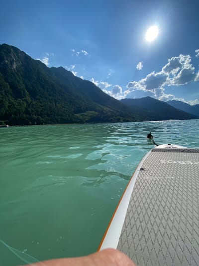 The media file shows a person stand-up paddleboarding on a lake. The sun is shining brightly in a blue sky with scattered clouds, suggesting it is daytime and the weather is clear. A paddleboard is visible in the foreground, with its textured surface and orange and white trim. The water is a calm, turquoise green. In the background, a lush, green mountain slopes down to the water's edge. The scene evokes a sense of peace and enjoyment of nature. The location appears to be a scenic body of water surrounded by mountains, consistent with a place like Lärchenwiese, Austria. No text is visible in the image.