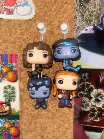 The image shows a corkboard with several items pinned to it. There are four Funko Pop figures of characters with oversized heads and simplified features pinned to the board. There are two blue figures, one brown-haired figure, and one red-haired figure. They appear to be attached with straight pins with round white heads. In the upper left corner is a portion of a photo showing grass and a dark-colored object. In the upper right corner is a portion of a patterned fabric or wallpaper with floral motifs. In the bottom left corner is a small section of a colorful cartoon-style image with a scalloped edge. In the bottom right corner is a fragment of a postcard or similar item with a vibrant floral design. The background is a plain cork surface. The image does not provide information regarding the city of Câmara de Lobos, Portugal.