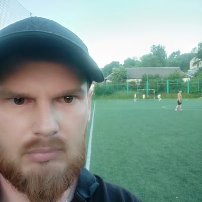 A man wearing a dark baseball cap and a dark shirt is in the foreground, taking a selfie on a bright, overcast day. His face, covered by a beard, is partially visible from the forehead down to his chin. In the background, a group of people are playing soccer on a green artificial turf field, marked with white lines. Trees and buildings are visible behind the field. The location is identified as Vyhoda, Ukraine.