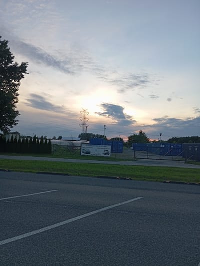 The image depicts an outdoor scene at dusk in Dybów Kolonia, Poland, featuring a road in the foreground and an industrial area with shipping containers in the background. The sky is a mix of blue and grey clouds, with the sun low on the horizon, casting a warm glow. A tall electricity pylon stands prominently in the middle ground.
In the foreground, there is an asphalt road with visible white lane markings. A curb separates the road from a grassy area.
The middle ground shows a fence surrounding an area filled with blue shipping containers. A white sign with Polish text is attached to the fence, advertising "WYPOŻYCZALNIA - WYNAJEM BUS-OSOBOWE DOSTAWCZE DO 3,5T" (Rental - Rental of Buses-Cars Delivery up to 3.5 tons). Below this text, a phone number is visible: "602 17 13 64". Adjacent to the fence, a row of dark green conical trees is planted.
The background consists of a line of trees against the horizon and more of the shipping containers. The overall impression is of an industrial site near a transportation route, with the natural elements of trees and sky creating a serene, yet functional, atmosphere. There are no people or animals visible in the image. The scene appears to be static, with no apparent activity or action.