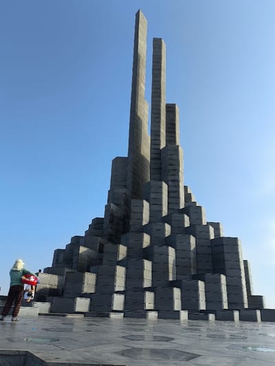 The image displays the Nghinh Phong Tower monument under a clear blue sky in Thành phố Tuy Hòa, Vietnam. The monument, constructed from dark grey, stacked, rectangular block-like components, rises from a wide, tiled plaza. Its base features a stepped, tapering design, culminating in multiple vertical pillars, with two prominent central pillars appearing vertically split near their apex. The ground is paved with dark grey hexagonal and rectangular tiles, featuring embedded circular clear panels that reflect light.
One individual is visible in the lower-left foreground, facing away from the camera, observing the monument. This person wears a light-colored hat, a light green long-sleeved shirt, and dark plaid trousers. They are holding a red and white inflatable ring. A second person, partially obscured behind the first, is visible in a white shirt. The scene is brightly lit by natural sunlight, casting subtle shadows.