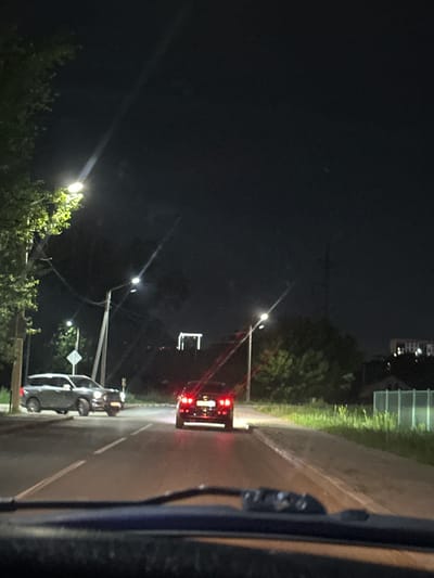 The image depicts a nighttime street scene, likely within the city of Kostanay, Kazakhstan. The perspective is from inside a vehicle, looking forward through the windshield. The road is paved and marked with white lane lines.
In the foreground, the top portion of the vehicle's dashboard is visible, out of focus.
On the road, two cars are visible. A darker colored SUV is in the left lane, facing away from the camera. To its right and further down the road, a darker sedan is also visible, facing away from the camera, its taillights illuminated.
Streetlights are present, casting pools of light on the road and illuminating the surrounding vegetation and trees. One streetlight is prominent on the left side of the image, with a visible beam of light. Another streetlight is visible further down the road on the right.
In the background, trees and vegetation line the sides of the road. Buildings and structures are faintly visible in the distance, suggesting an urban or suburban environment. A tall, rectangular structure, possibly a building or pylon, is illuminated by lights in the middle distance. A fence, possibly a green chain-link fence, is visible on the right side of the road in the background. The sky is dark, indicating nighttime.