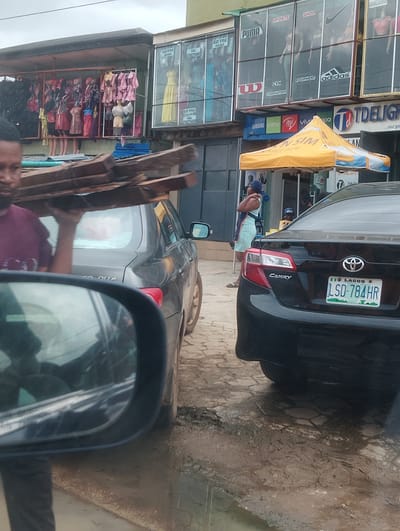A medium shot captures an outdoor market scene in Iju, Nigeria. In the foreground, a man in a maroon t-shirt carries a stack of wooden planks on his head, walking past a gray car. To the right, a black Toyota Camry is parked, displaying a Nigerian license plate that reads "LSD-784HR". Behind the car, a yellow tent is set up. The background reveals a row of market stalls with various goods, including clothing displayed on mannequins, and shop fronts with advertising signs for brands like "Puma", "Adidas", and "Samsung". A woman in a light blue dress is visible near the tent. The ground is uneven and appears to be a mixture of paved and muddy areas. The sky is overcast.