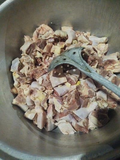 In Sapele, Nigeria, a close-up shot reveals a large, stainless steel bowl filled with raw, chopped chicken pieces. A light gray, slotted serving spoon rests on top of the chicken. The chicken pieces vary in size, with some showing skin and others displaying exposed meat and bone. The bowl appears to be in a kitchen setting, judging by the metallic sheen and the food preparation context. The lighting is dim, casting shadows and highlighting the texture of the meat and the surface of the bowl.