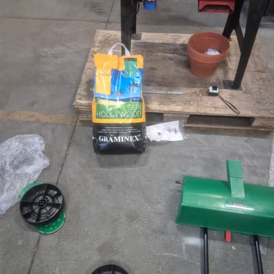 The image depicts a scene on a concrete floor, possibly in a storage area or workshop, in Nowa Wieś Książęca, Poland. In the center of the image, there is a large yellow and blue bag of grass seed labeled "SUN HOLLYWOOD" and "GRAMINEX". The bag is positioned on a wooden pallet. To the right of the bag, there is an orange clay pot containing some white material. A small black and white measuring tape is also visible on the pallet. In the lower left corner, a black wheel with green accents is placed on the ground. To the right and slightly further back, a green gardening tool with black legs is visible. A crumpled plastic bag is partially visible to the left of the seed bag. The environment is utilitarian, with a concrete floor and industrial elements like a metal frame supporting some equipment in the background. There are no people visible in the image.