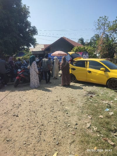 People gather near a yellow car with an open trunk, under a reddish umbrella. Several motorcycles are parked alongside them. Most of the people are wearing long dresses or robes and head coverings, with one man in a plaid shirt. A building and trees are in the background. The ground is dirt and grass with some trash. A date and time are visible in the bottom right corner of the image.